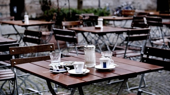 Café-Terrasse ohne Gäste, mit zwei Kaffeetassen und Wassergläsern an einem leeren Tisch