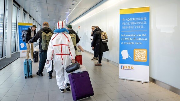 Reisende aus China gehen auf einem Flughafen an einem Banner mit Informationen zu Corona-Tests vorbei