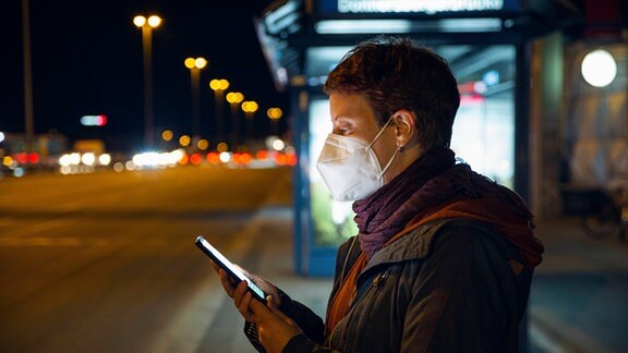 Eine Frau trägt eine FFP2-Maske, schaut auf ihr Handy und wartet an einer Bushaltestellte