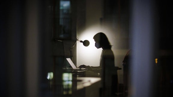 Eine Frau sitzt abends in ihrem Schreibtisch im Buero und traegt eine FFP2 Maske.