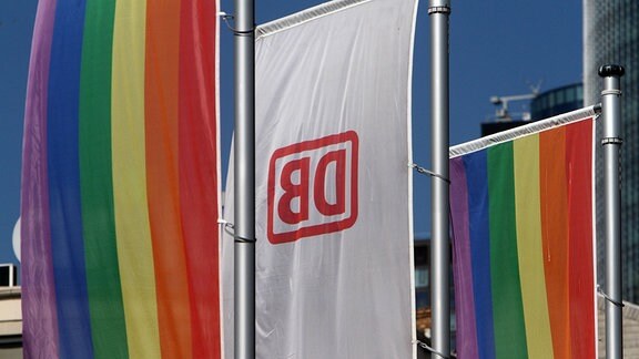 Eine Flagge mit dem Logo der DB Deutsche n Bahn wird in Frankfurt von zwei Regenbogenflaggen eingerahmt,