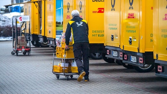 Ein Mitarbeiter der Deutschen Post schiebt einen Wagen mit Paketen hinter einer Reihe von DHL-Transportern