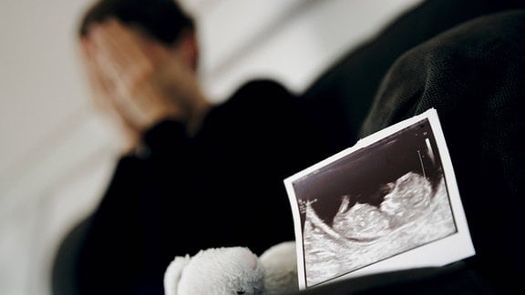 Symbolfoto: Gestellte Aufnahme zum Thema Fehlgeburt. Das Ultraschallbild eines ungeborenen Kindes steht neben einem Kuscheltier waehrend eine Frau im Hintergrund ihr Gesicht in ihren Händen verbirgt. 