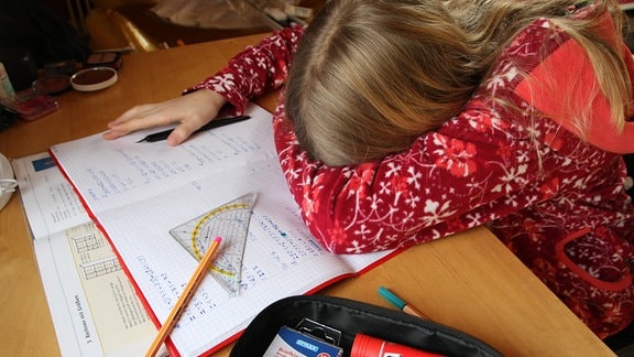 Ein Mädchen sitzt am vollen Schreibtisch und versteckt ihr Gesicht im gebeugten Arm