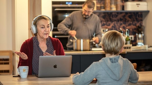Frau im Homeoffice mit Kopfhörern am Laptop, ihr Mann kocht währenddessen, ihr Enkel macht Hausaufgaben