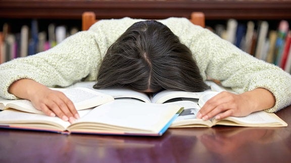 Eine junge Frau, die in der Bibliothek eines Colleges lernt und mit ihrem Gesicht auf den Büchern liegt.