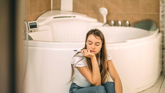 Inszeniertes Foto einer Traurige Frau, die einen Schwangerschaftstest hält  und im Bad auf dem Boden sitzt.