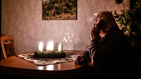 Ein Mann sitzt am Tisch und telefoniert. Auf dem Tisch brennen drei von vier Kerzen.