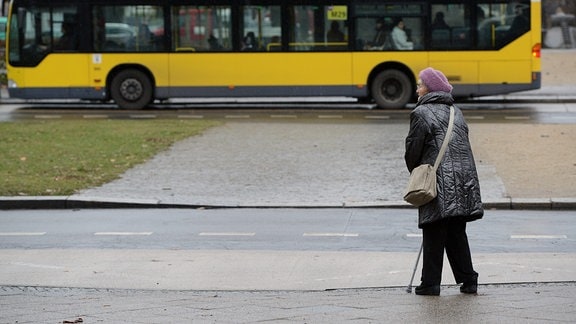 Eine Seniorin steht am Straßenrand, auf der anderen Straßenseite hält ein Bus