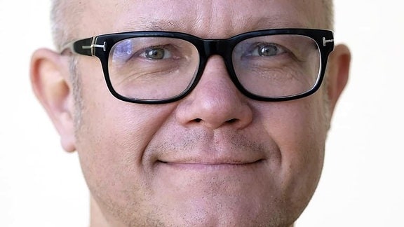 ein Mann mit Glatze und Brille lächelt vor weißem Hintergrund in die Kamera