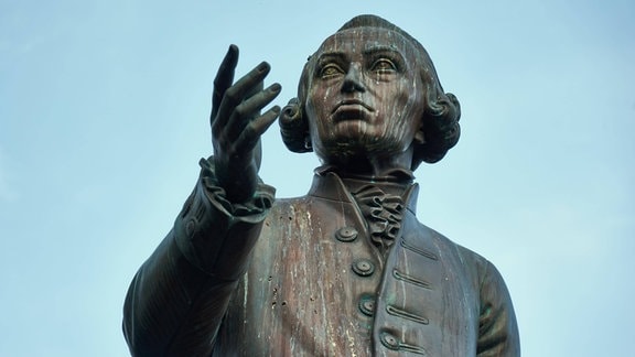 Statue eines Mannes mit Perücke, er hält gestikulierend eine Hand vor sein Gesicht.