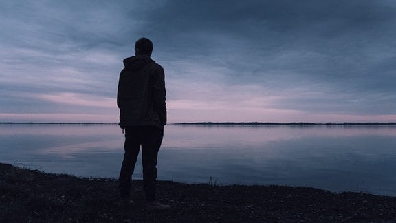 Einsamer junger Mann steht an Seeufer und blickt in die Ferne