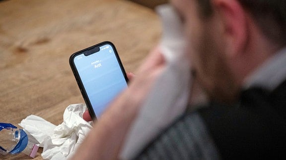 Ein Mann hält ein Taschentuch in einer Hand und ein Telefon mit dem Schriftzug «Arzt» in der Anderen.