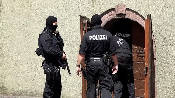 Drei vermummte Polozisten mit Gewehren, zwei stehen vor einer Hauswand, der dritte betritt das Gebäude durch eine Tür.