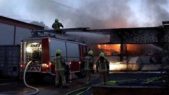 Eine Halle, in der Flammen lodern. Rundrum Feuerwehr im Löscheinsatz