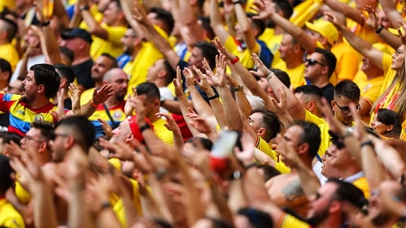 Zahlreiche gelb gekleidete Menschen, rumänische Fans, auf einer Tribüne strecken ihre Arme in die Höhe.