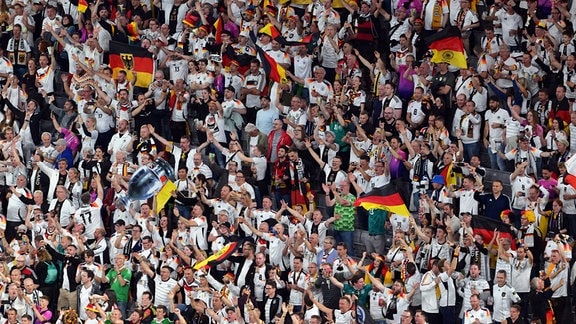 Eine Menschenmasse in der Fußball Arena München mit Deutschlandfahnen und anderen schwarz-rot-goldenen Fanartikeln reißt freudig die Arme in die Luft.