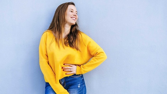 Eine junge Frau mit langen Haaren und gelben Pullover schaut nach recht sund lächelt.
