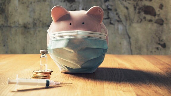 Sparschwein trägt medizinische Schutzmaske mit Goldmünzen und einer Spritze Impfstoff