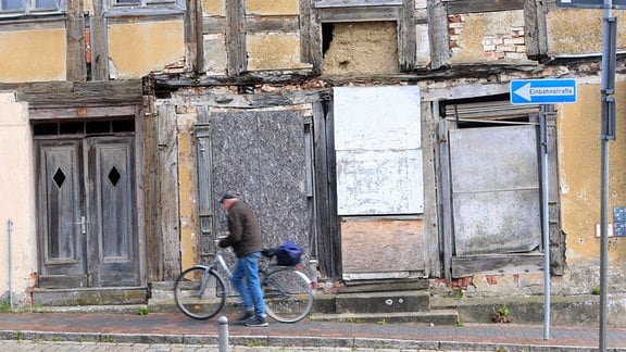 Älterer Mann schiebt vor leerstehenden Gebäuden ein Fahrrad eine verlassene Straße entlang
