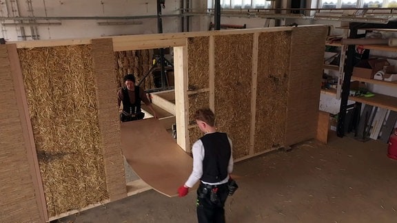 Zwei Handwerker arbeiten in der Halle an einem Strohballen Modul.