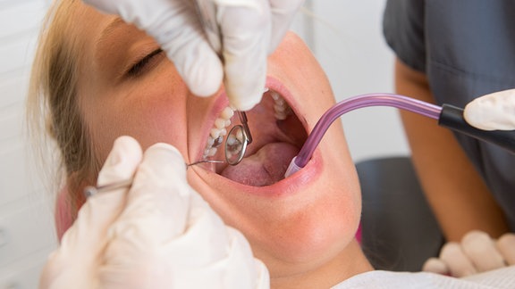 Ein Zahnarzt begutachtet die Zähne einer Frau, während eine Zahnarzthelferin mit einem Absauger assistiert.