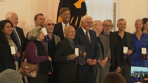 Alle Preisträger:innen des Bundesverdienstkreuzes stehen um Bundespräsident Steinmeier. Zu den bekannten Gesichtern gehören der City-Sänger Toni Krahl und Fußballtrainer Jürgen Klopp