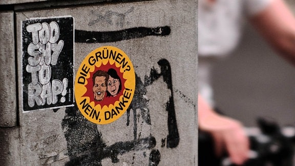 An einem Verteilerkasten klebt ein Aufkleber, dem Logo der Antiatomkraftbewegung nachempfunden, mit dem Schriftzug "Die Grünen? Nein Danke!" und Karikaturen der Parteivorsitzenden von Bündnis90/Die Grünen, Habeck und Baerbock. 