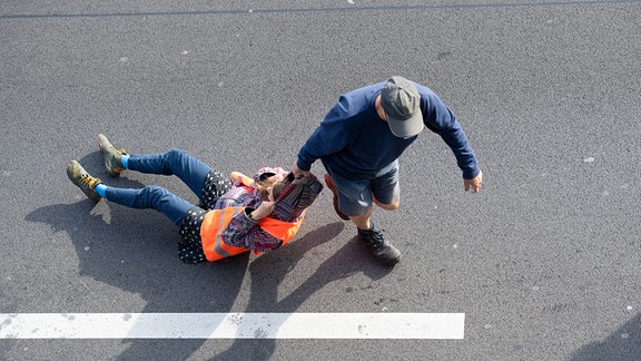 Ein Verkehrsteilnehmer (r) zieht bei einer Blockade der Letzten Generation auf der Autobahn 100 eine Aktivistin von der Straße. 