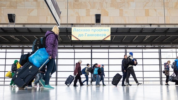 Geflüchtete aus der Ukraine laufen nach ihrer Ankunft durch die Eingangshalle vom Messebahnhof Laatzen. Auf einem Banner im Hintergrund steht «Willkommen in Hannover» auf Ukrainisch.