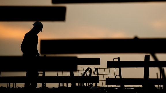 Die Silhouette eines Bauarbeiters zeichnet sich auf einer Baustelle vor dem verfärbten Morgenhimmel ab
