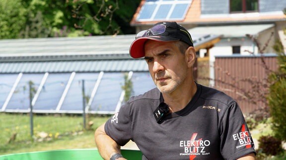 Ein mann in einer Interviewsituation. Er sitzt vor einem Haus mit Solarpanellen. 