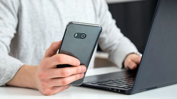 ILLUSTRATION - Ein junger Mann sitzt im Homeoffice am Laptop und hält ein Smartphone in der Hand.