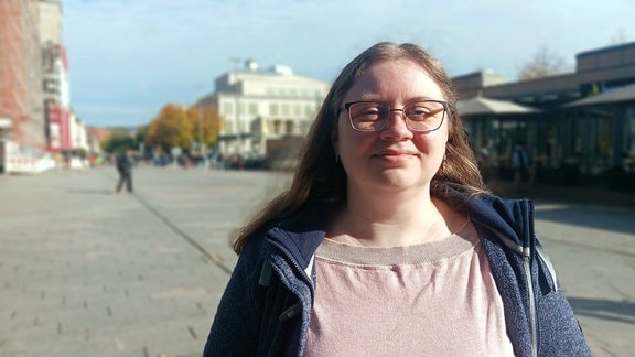 Eine Frau mit braunen langen Haaren und einer Brille steht in Leipzig auf einem asphaltierten Platz. Verschwommen im Hintergrund sieht man die Oper Leipzig. 