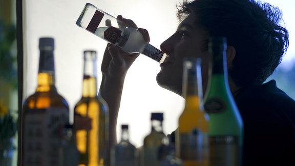 Ein Jugendlicher sitzt hinter Flaschen mit Alkohol und trinkt ein alkoholhaltiges Mixgetränk