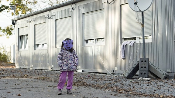 Ein Mädchen mit einer Maske steht vor einer Reihe Containern