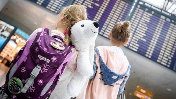 Zwei Mädchen stehen vor einer Abflugtafel an einem Flughafen
