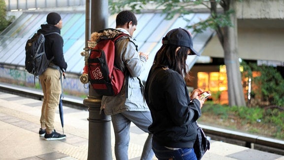 Junge Leute auf einem Bahnsteig haben ihre Häupter gen Smartphone gesenkt.