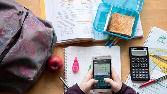 Eine Schülerin sitzt mit ihrem Handy und ihren Schulsachen an einem Schreibtisch. 