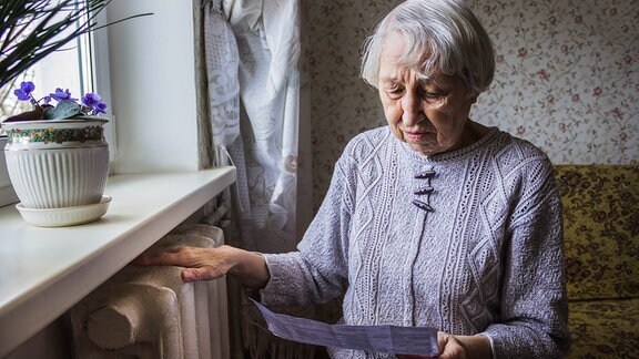 Seniorin liest eine Rechnung, während sie eine Hand auf die Heizung hält.