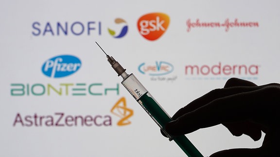 Eine Spritze vor den Logos der Unternehmen Sanofi, Biontech, Pfizer, AstraZeneca, Curevac, Johnson & Johnson, Sanofi und GlaxoSmithKline