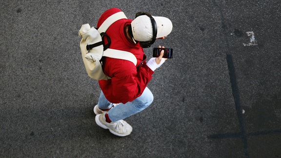Ein junger Mann trägt Kopfhörer und blickt auf sein Smartphone, während er in Steglitz über eine Strasse geht. 