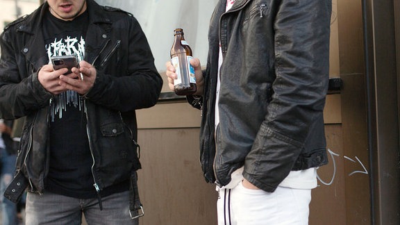 Zwei jüngere Männer stehen auf der Straße mit Smartphone und Bierflasche in der Hand.