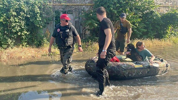 Helfer ziehen einen Mann und einen Hund in einem Gummiboot durch überschwemmtes Gebiet.