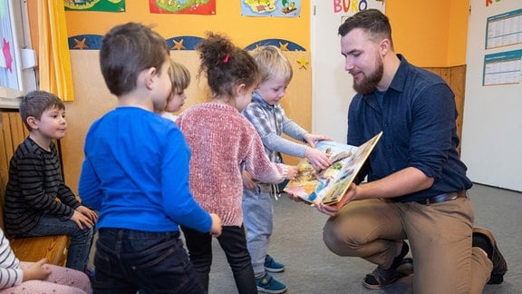 Ein Erzieher im Kindergarten bespricht mit Kindern zur musikalischen Früherziehung ein Buch über Zootiere.