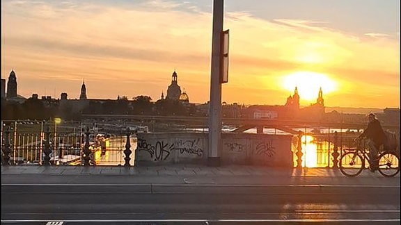 Ein*e Radfahrer*in überquert während eines Sonnenuntergangs eine Brücke.