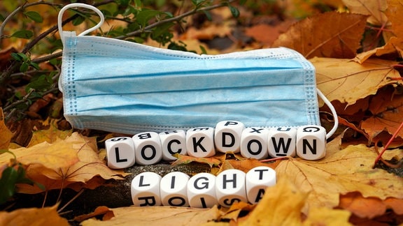 Ein blauer Mundschutz liegt auf Herbstlaub, davor bilden Würfel das Wort: Lockdown Light