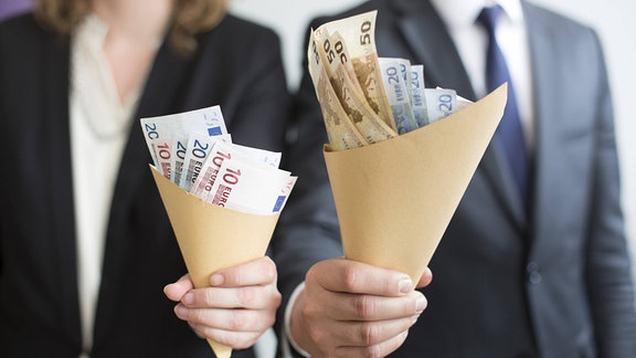 Mann und Frau halten unterschiedlich große Papiertüten gefüllt mit Geldscheinen in den Händen.