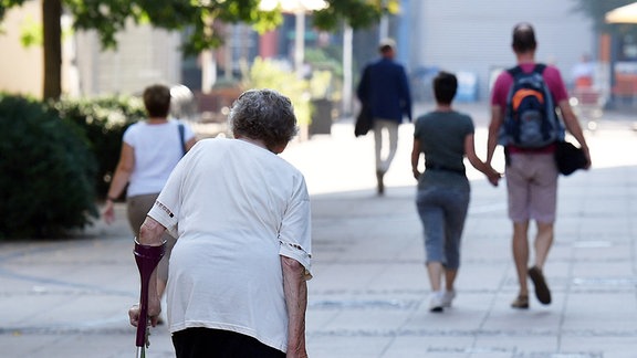Eine alte Frau mit einer Krücke und Einkaufstrolley sowie ein junges Paar gehen in Magdeburg in der Fußgängerzone.