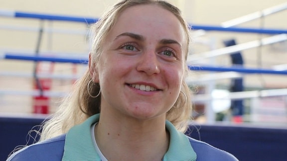 Junge Frau schaut lächelnd in die Kamera, im Hintergrund ein Boxring.